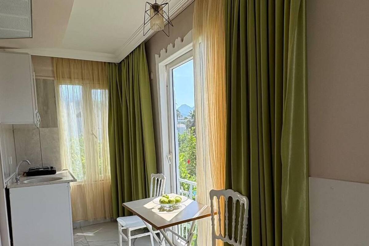 Une pièce de taille modeste comprenant un lit, une petite table avec des chaises, un coin cuisine et une vue sur la verdure environnante.