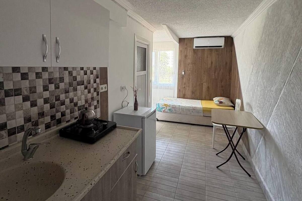 Une cuisine compacte équipée d’un évier, d’une cuisinière et de placards. Une petite table et un banc se trouvent dans la pièce adjacente.