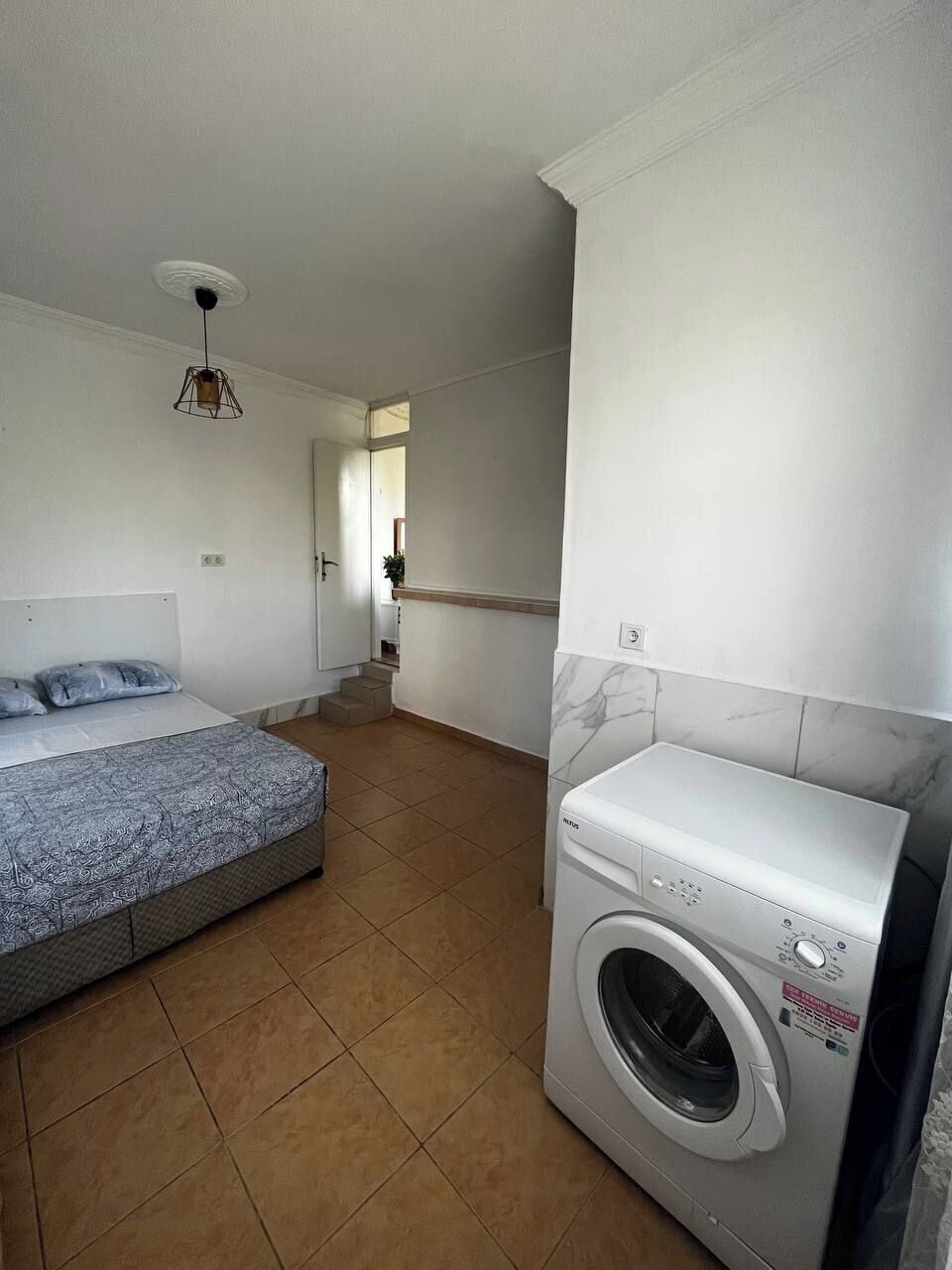 A small, clean room with a bed, a washing machine, and a staircase leading to another area.
