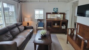 Een woonkamer met een bruine leren bank, een houten salontafel en een televisie aan de rechterkant. Er is een raam met uitzicht, een lamp en een houten kast met een schilderij.