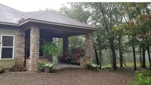 A house with a stone porch, a window with shutters, and a small garden area with plants and a fire pit.