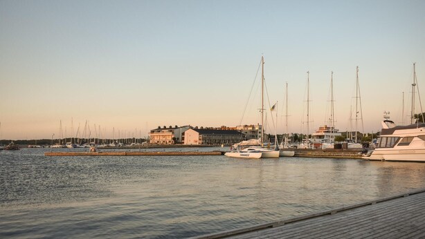 A marina with several yachts docked, a wooden pier, and buildings in the background.