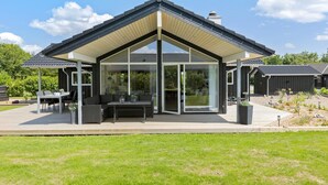 Una casa moderna con una amplia puerta de vidrio, un porche cubierto con mobiliario exterior y un jardín bien cuidado.