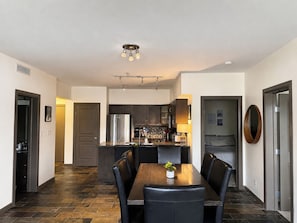 A modern kitchen with dark cabinetry, a stainless steel refrigerator, and a dining area with a wooden table and black chairs.