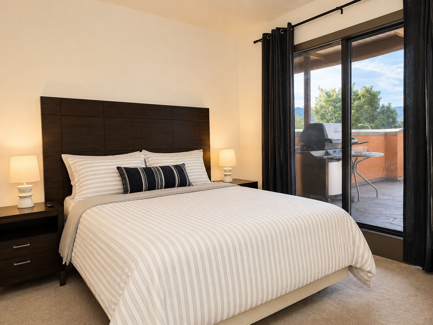 A hotel room with a large bed, two bedside tables with lamps, a view of the outdoors through a sliding door, and a patio area with a grill.