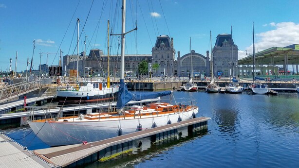 Un puerto deportivo con barcos amarrados, un edificio grande con una cúpula al fondo y una zona cubierta con gente.