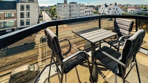 Un balcón con una mesa y sillas, que ofrece vistas a un paisaje urbano con edificios y una vía ferroviaria.
