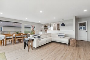 Uma sala de estar moderna com um sofá branco, cadeiras de madeira, uma mesa de jantar e uma área de cozinha ao fundo.