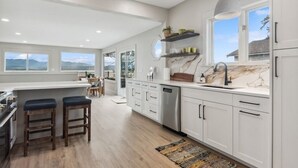 A modern kitchen with white cabinets, a marble countertop, and a large window offering a view of the mountains.