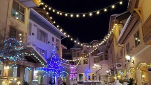 A snow-covered street with buildings on either side, decorated with string lights and illuminated trees. - Spacious Alpine Retreat - w/Hot Tub (Sun Peaks)