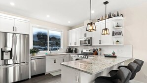 A modern kitchen with white cabinetry, stainless steel appliances, a large island with bar stools, and pendant lighting. - Spacious Alpine Retreat - w/Hot Tub (Sun Peaks)