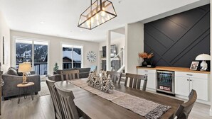 A modern living room with a dining area, large windows with a view of mountains, a kitchen with a black accent wall, and a staircase. - Spacious Alpine Retreat - w/Hot Tub (Sun Peaks)