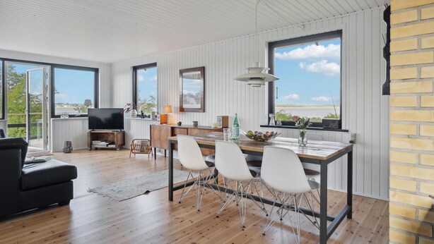 A spacious living area with a wooden floor, a dining table with chairs, a TV, and a large window with a view of the sea.