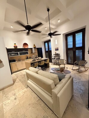 A modern living room with a sofa, a dining area with chairs, a kitchen with cabinets, and a ceiling fan.