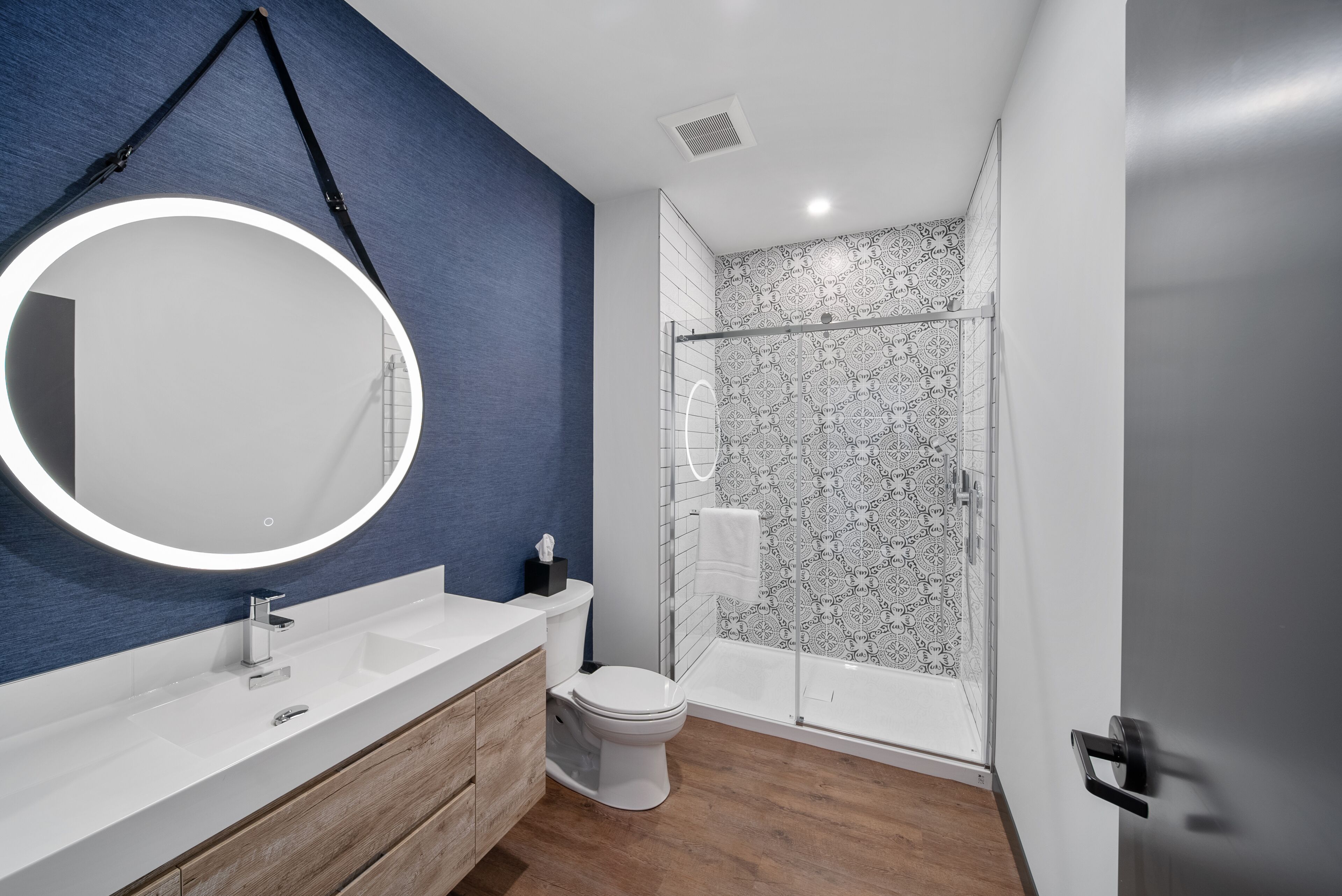 A modern bathroom with a round mirror, a white sink, a wooden vanity, a toilet, and a glass-enclosed shower.