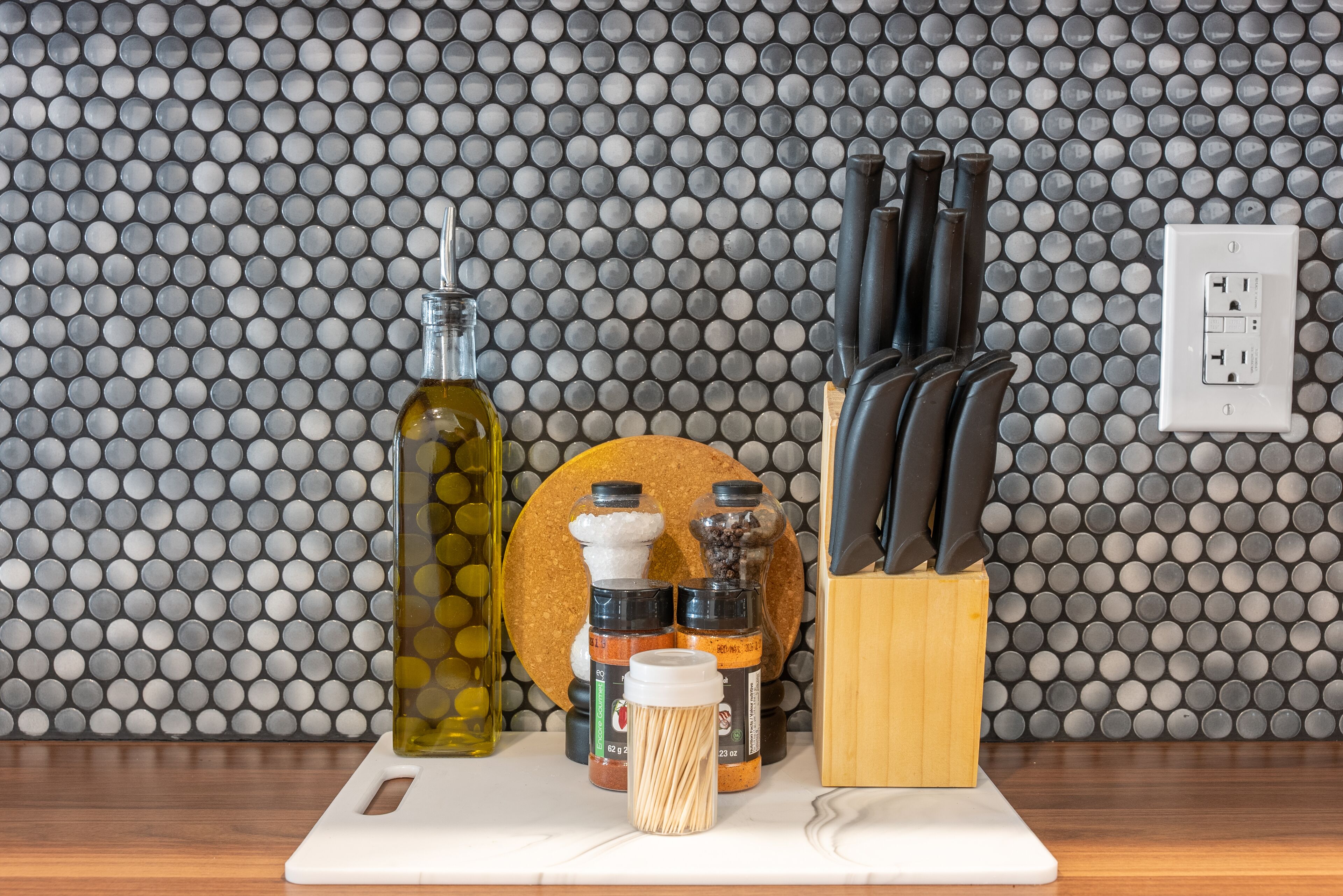 Una encimera de cocina con tabla de cortar, una botella de aceite de oliva, frascos de especias y un bloque de cuchillos.