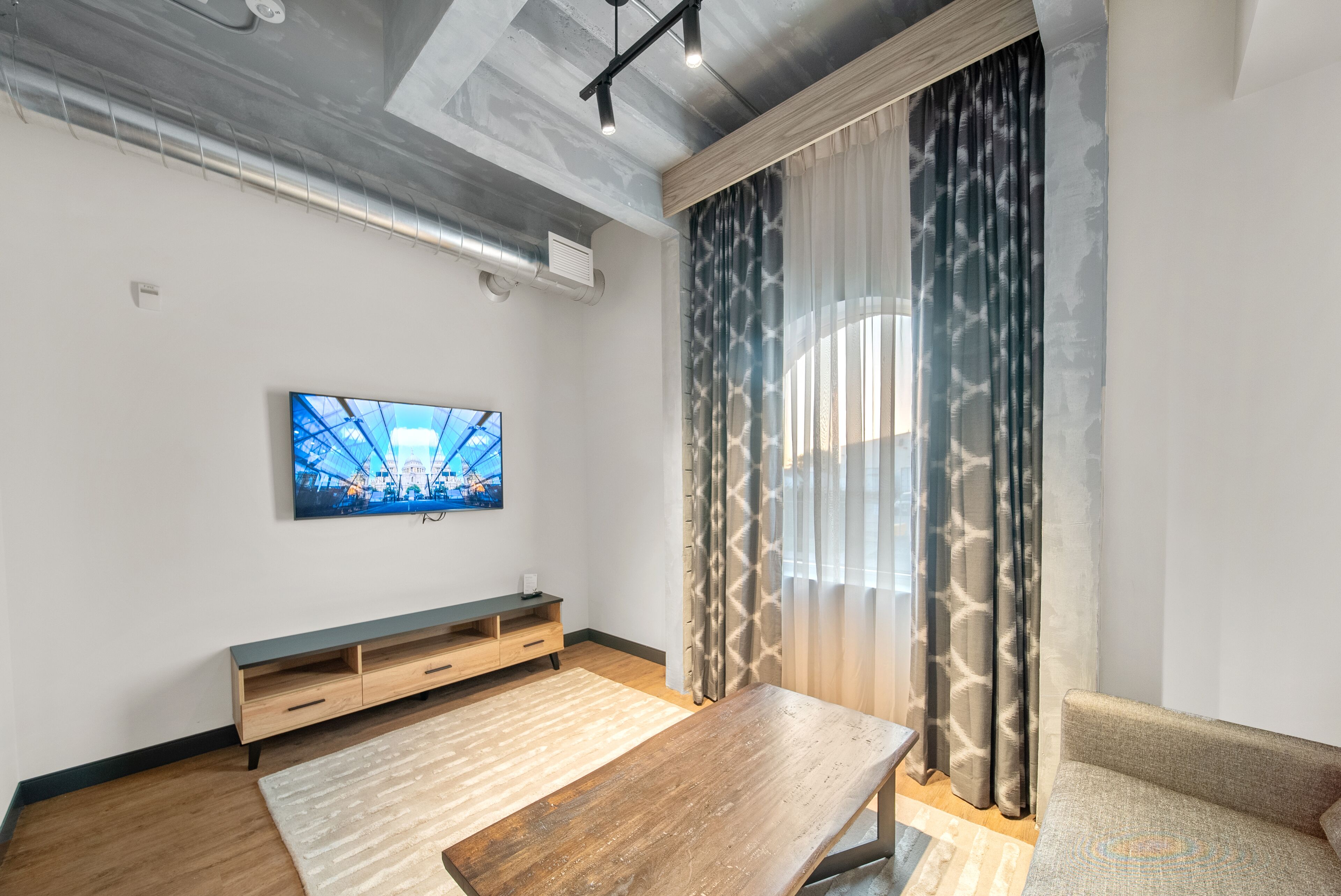 A modern living room with a wooden coffee table, a sofa, a TV mounted on the wall, and patterned curtains.
