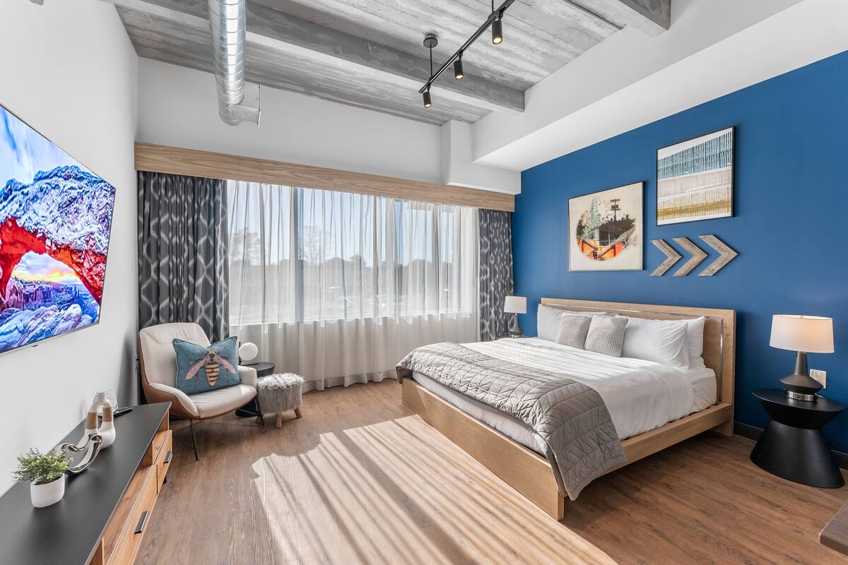 A modern bedroom with a large bed, a desk, and a chair. There are two paintings on the wall and a window with curtains.