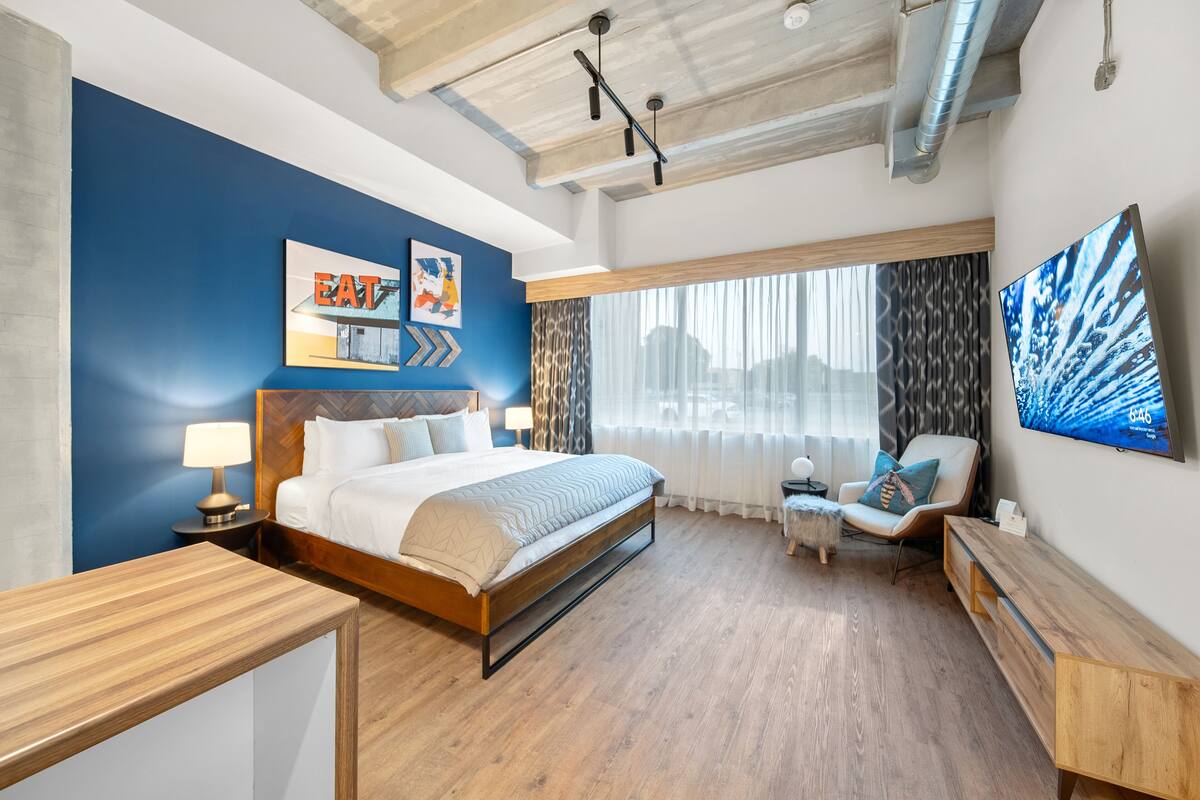 A modern hotel room with a large bed, a wooden nightstand, a chair, a TV, and a window with curtains.