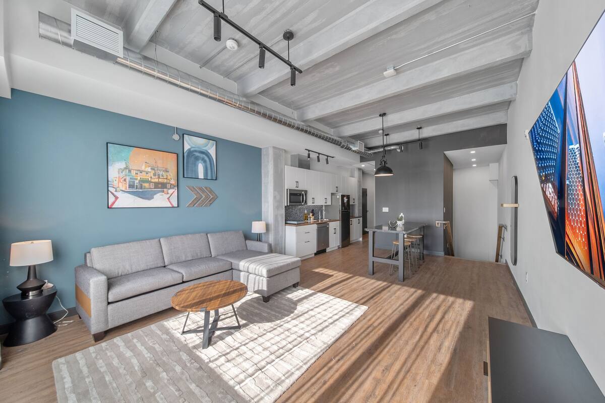 A modern living room with a grey sofa, a wooden coffee table, and a striped rug. There is a kitchen area with white cabinets and a dining table.