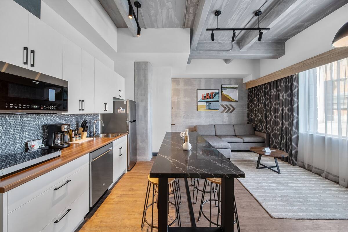 A modern kitchen with white cabinets, a black countertop, and a marble island. There is a dining area with a sofa and a coffee table.