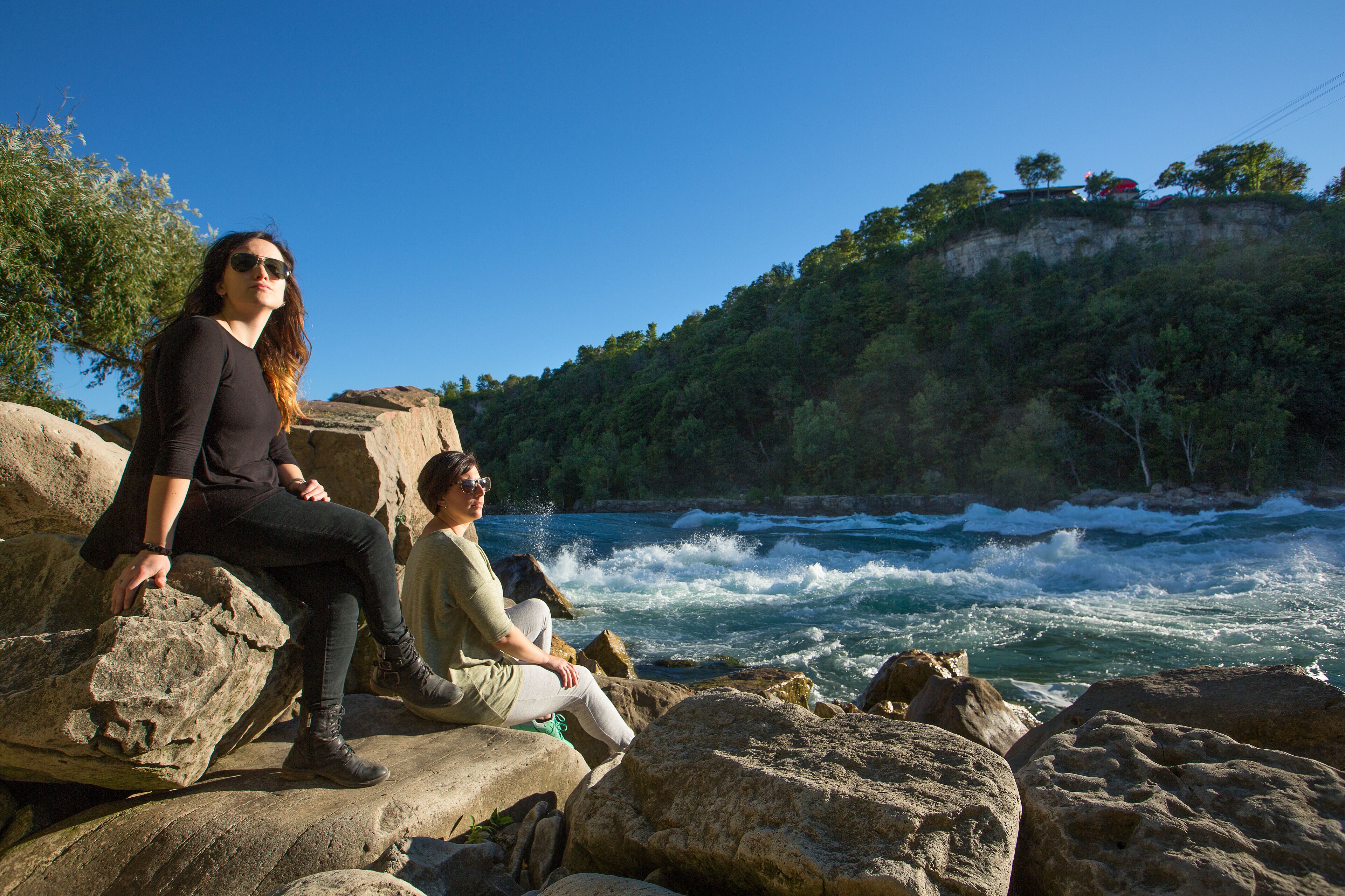 Duas pessoas sentadas em pedras grandes perto de um rio, com uma colina arborizada ao fundo.
