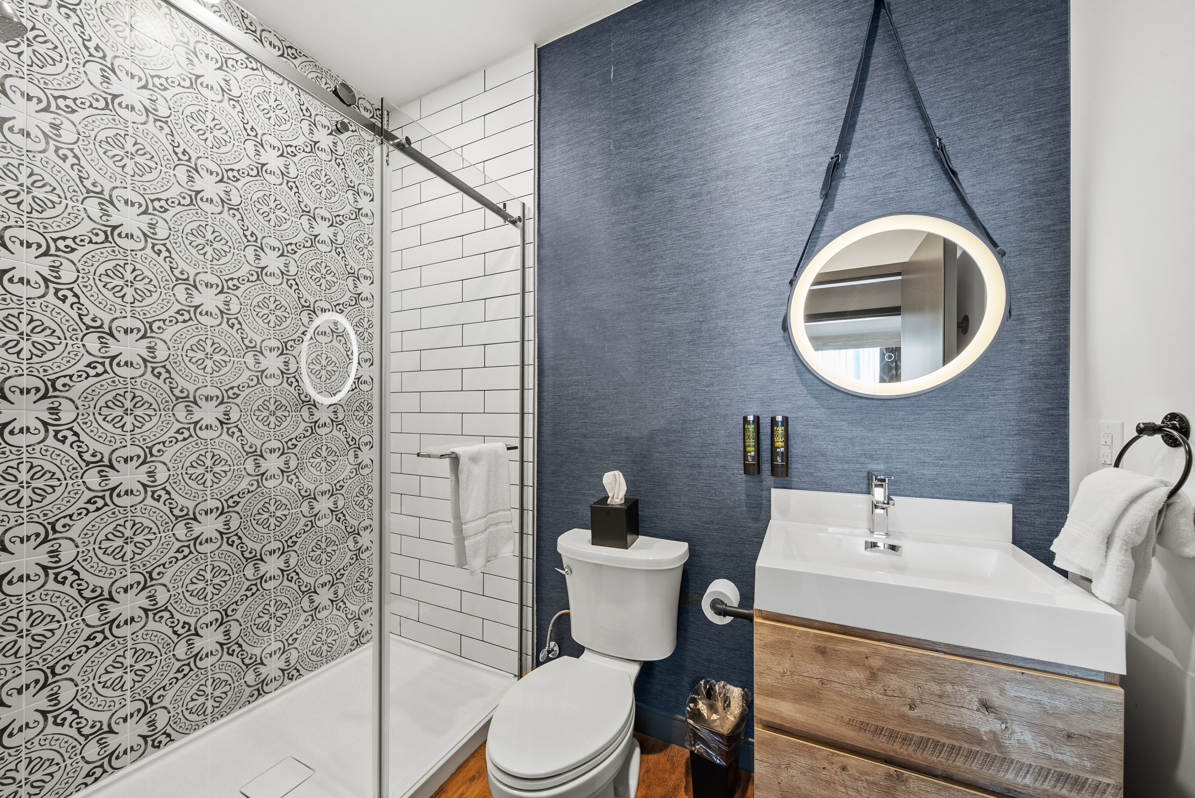 A bathroom with a patterned shower wall, a toilet with a tissue box, a sink with a mirror, and a wooden vanity.