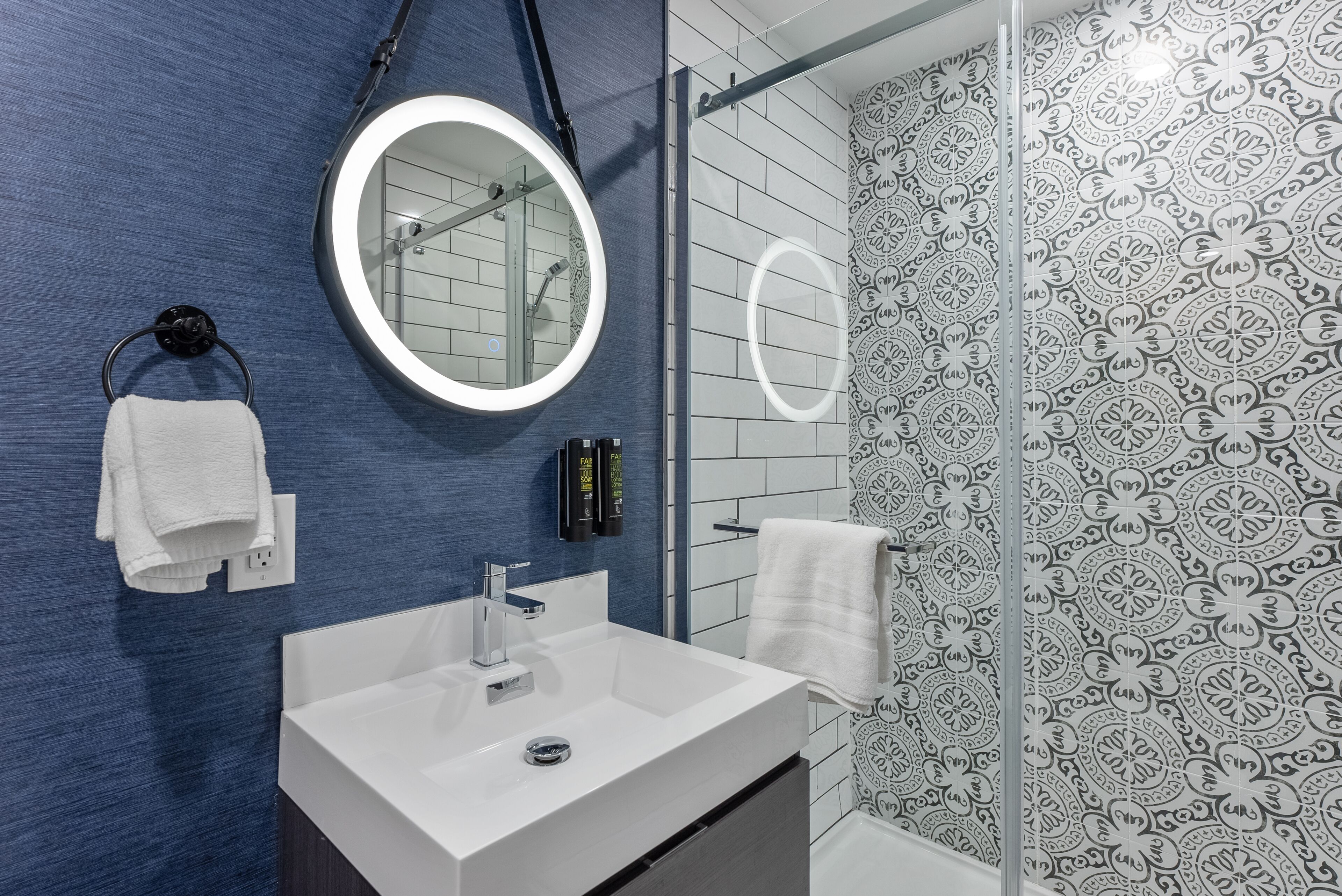 Ein modernes Badezimmer mit blauer Wand, Waschbecken in Weiß, rundem Spiegel und Dusche mit gemusterten Fliesen.