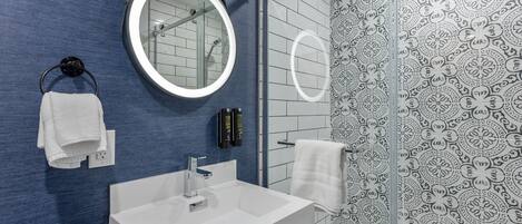 Ein modernes Badezimmer mit blauer Wand, Waschbecken in Weiß, rundem Spiegel und Dusche mit gemusterten Fliesen.