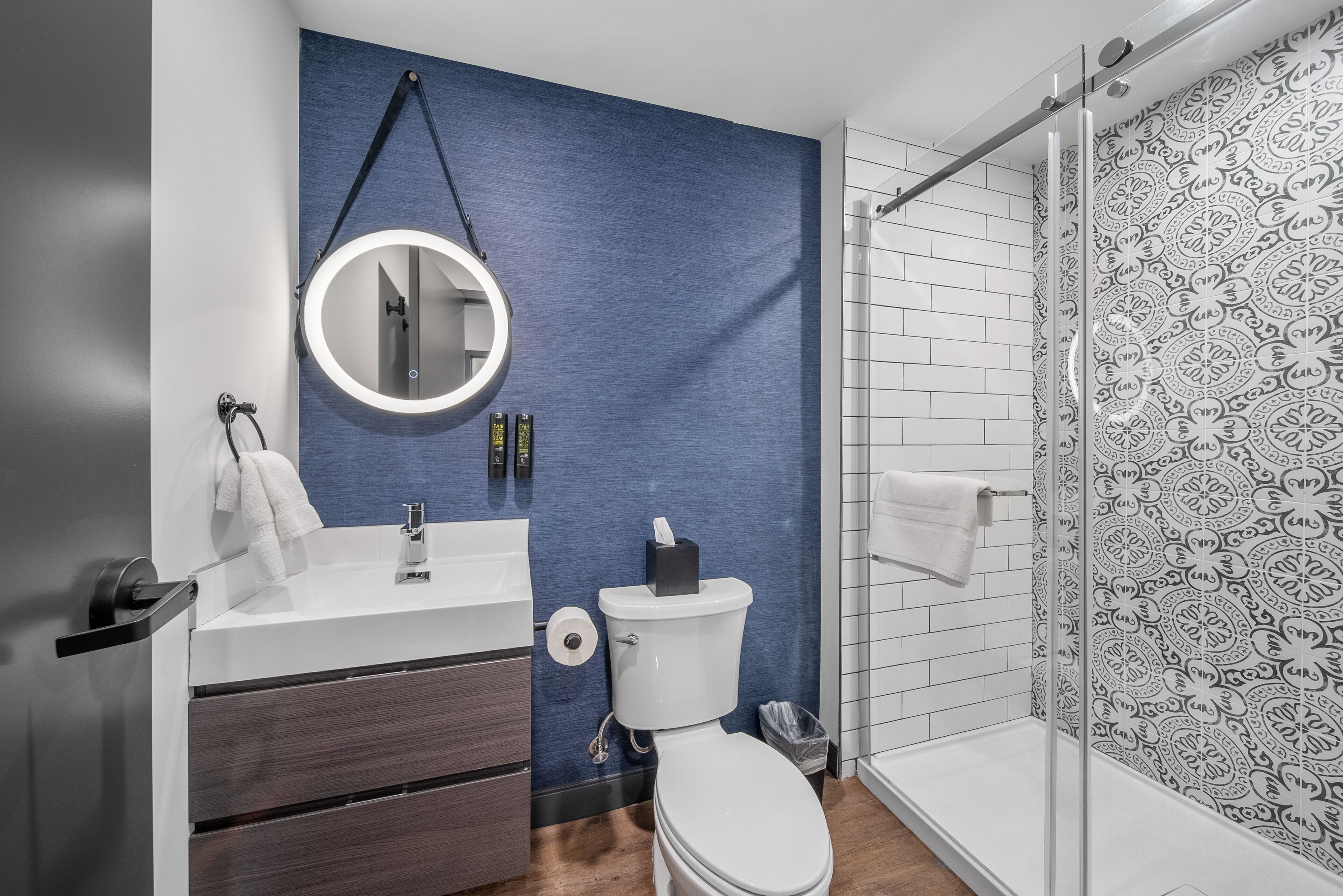 A modern bathroom with a blue accent wall, a round mirror, a white sink, and a glass shower enclosure.