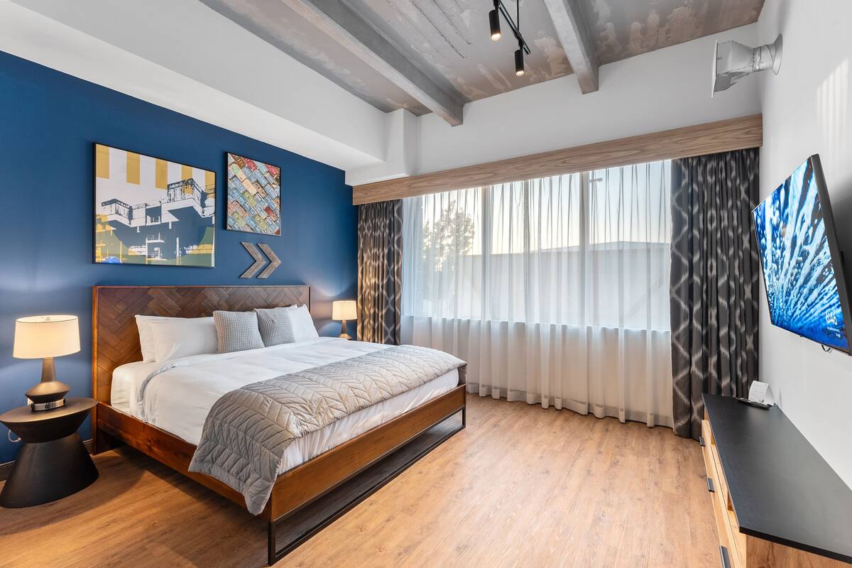 A modern bedroom with a wooden bed, a nightstand, a desk, and a large window with curtains.