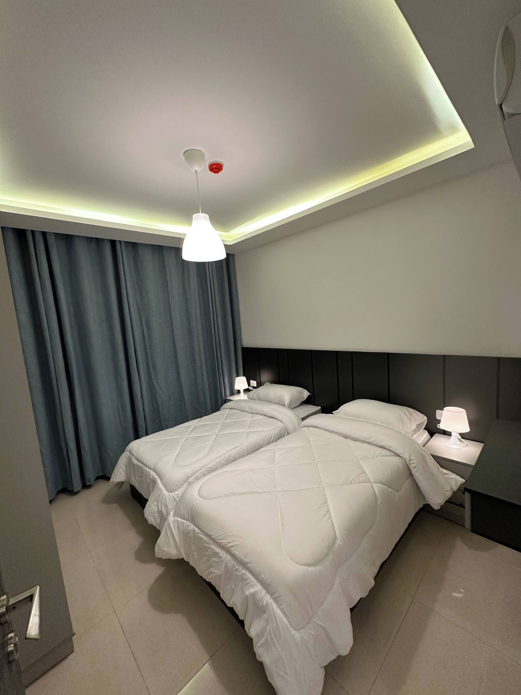 Quarto de hotel com cama de casal, mesa de cabeceira com luminária, luz no teto e janela com cortinas.
