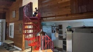 Interior de una cabaña de madera con piso de cuadros, una escalera espiral roja, un área de cocina con refrigerador, y una persona sentada en la escalera.
