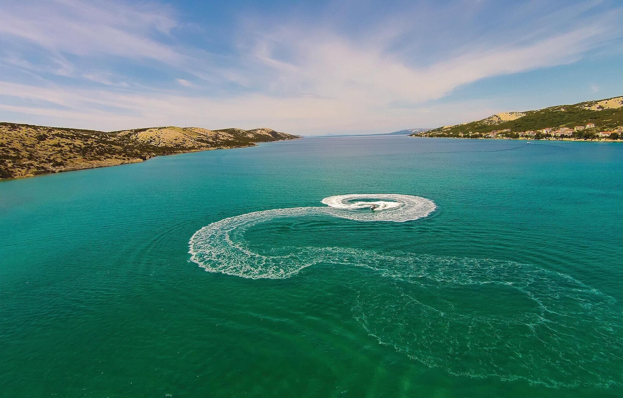 An aerial view of a large body of water with a circular pattern on the surface, surrounded by hills and a distant shoreline with buildings.