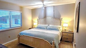 Une chambre à coucher avec un grand lit, deux tables de chevet, une fenêtre avec des stores et un luminaire au plafond.