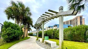 Un chemin menant à un bâtiment agrémenté d’une pergola et de bancs, entouré de verdure et de palmiers.