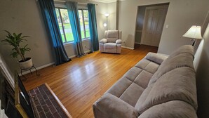 A living room with a beige sofa, a chair, a potted plant, and a window with curtains. - Gorgeous model single family home, fully renovated and ready for you.  (Newport News)