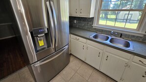 A stainless steel refrigerator with a digital display, a built-in ice maker, and a freezer compartment. The kitchen has white cabinets, a granite countertop, and a window with a view of the outdoors. - Gorgeous model single family home, fully renovated and ready for you.  (Newport News)