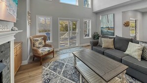 A spacious living room with a fireplace, a large sofa, a coffee table, and a view of the deck through the windows. - Renovated Cottage w/ Heated Pool, Spa & Amenities! (Corolla)