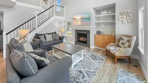 A spacious living room with a fireplace, a staircase, and a view of the beach. - Renovated Cottage w/ Heated Pool, Spa & Amenities! (Corolla)
