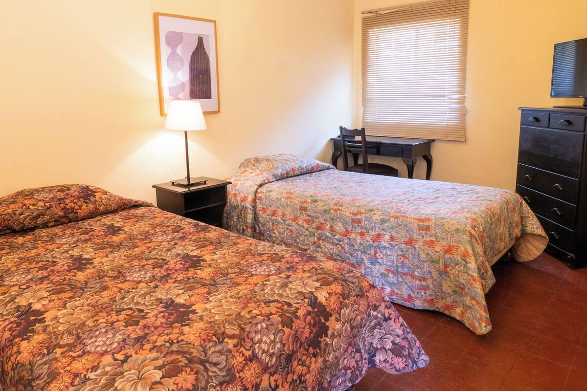 Une chambre avec deux lits, un bureau, une chaise, une lampe, une commode et une fenêtre avec des stores.