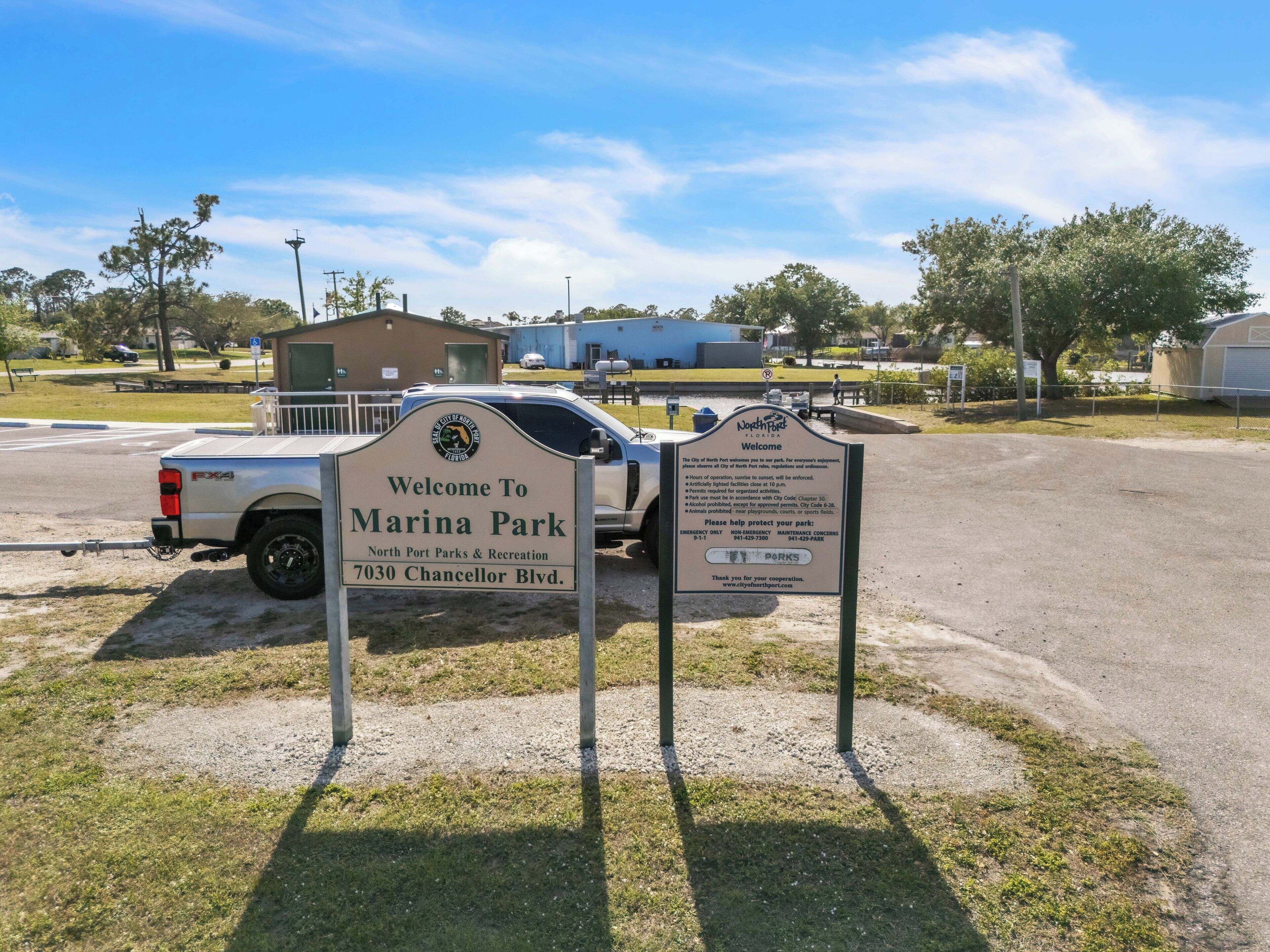 Un panneau d’accueil à l’entrée du parc Marina, géré par le service des parcs et des loisirs de North Port, avec un espace de stationnement et un bâtiment en arrière-plan.
