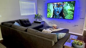Un salon moderne avec un canapé d’angle, un téléviseur à écran plat fixé à un mur en briques, une petite table d’appoint avec une plante et un seau à pop-corn.