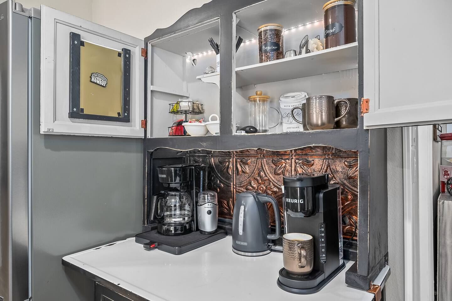 Une petite cuisine équipée d’une machine à café, d’une bouilloire et d’une tasse sur le plan de travail, ainsi que d’étagères au-dessus contenant des bocaux et des ustensiles de cuisine.