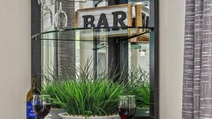 Un coin bar en bois sombre, avec une étagère en verre où sont disposés des verres à vin et des bouteilles, une plante en pot et un panneau indiquant « BAR ».