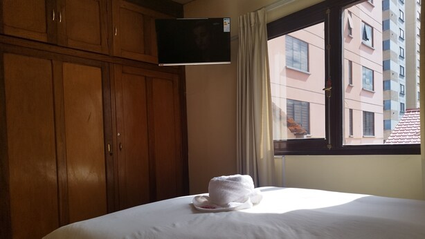 A hotel room with a bed, a TV mounted on the wall, and a window with a view of buildings. - Hotel RustiCall (La Paz)