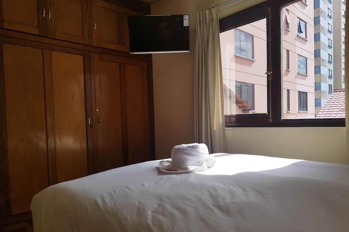 A hotel room with a bed, a TV mounted on the wall, and a window with a view of buildings.