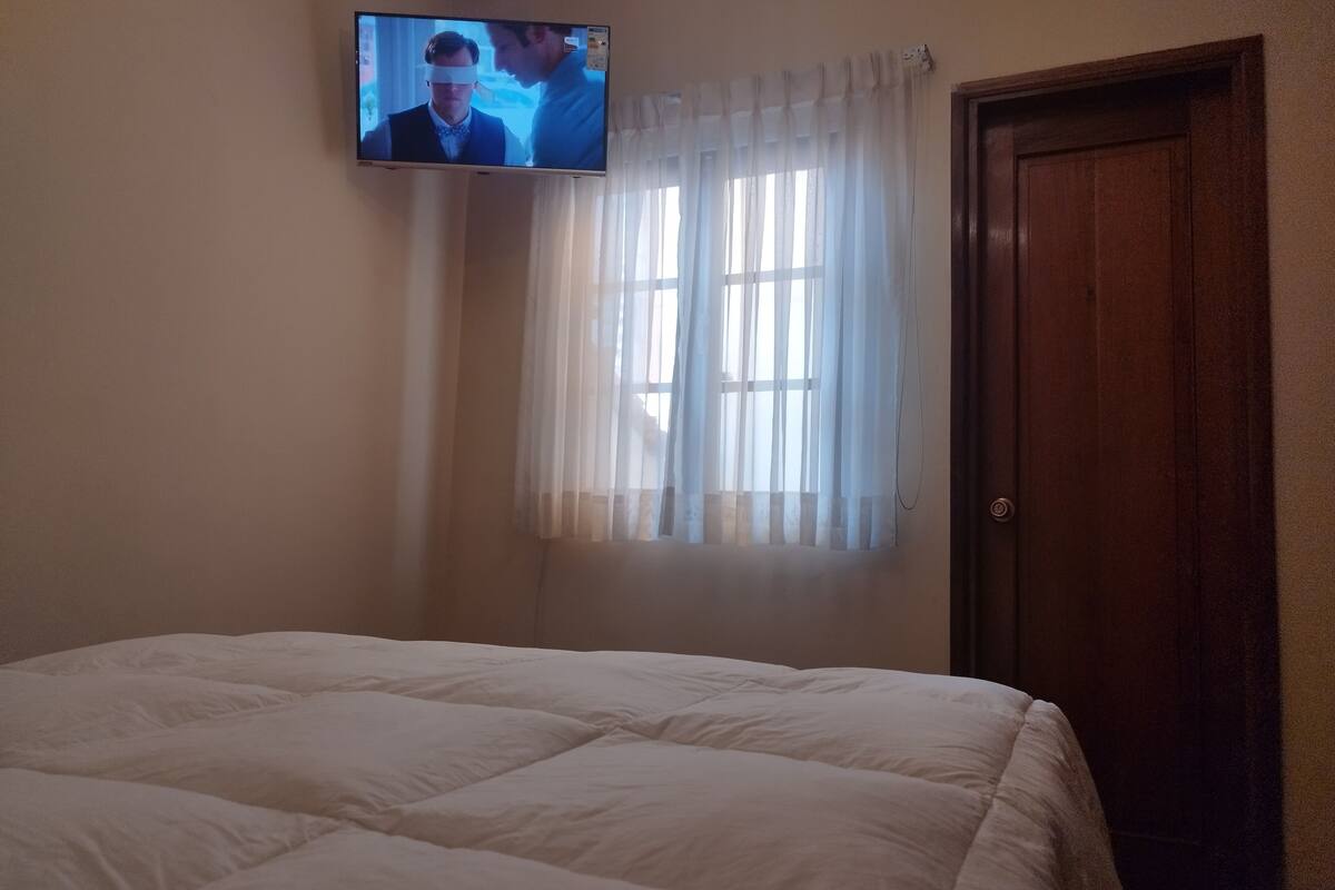 A bedroom with a bed, a television mounted on the wall, a window with sheer curtains, and a wooden door.