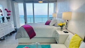 Una habitación de hotel moderna con balcón, una cama con colcha blanco y rosa, un sofá blanco, una mesa de centro de cristal y una alfombra color turquesa.