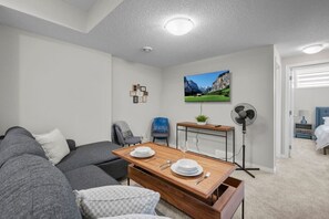 Una sala de estar moderna con un sofá, una mesa de comedor dispuesta con platos y vasos, una silla, un mueble pequeño, un ventilador y un cuadro en la pared.